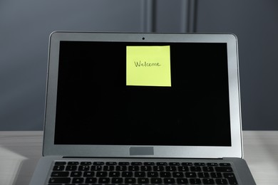 Photo of Sticky note with word Welcome and laptop on white wooden table against grey background, closeup