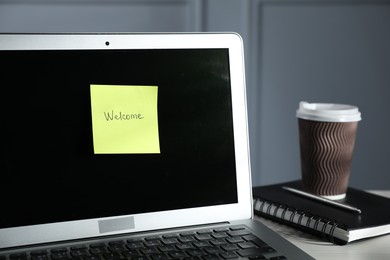 Photo of Sticky note with word Welcome, laptop, notepad and takeaway cup on light wooden table against grey background, closeup