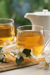 Photo of Tasty jasmine tea in cups, teapot and flowers at white wooden table, closeup