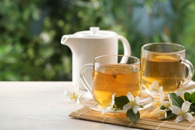 Photo of Tasty jasmine tea in cups, teapot and flowers at white wooden table, closeup. Space for text