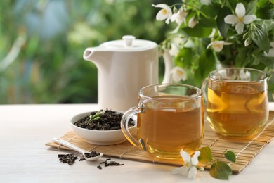 Photo of Tasty jasmine tea in cups, teapot, dry leaves and flowers at white wooden table, closeup. Space for text