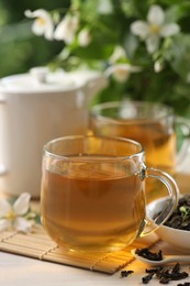 Photo of Tasty jasmine tea in cups, teapot, dry leaves and flowers at white wooden table, closeup