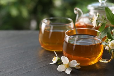 Photo of Tasty jasmine tea in cups, teapot and flowers on black table, closeup. Space for text