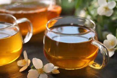 Photo of Tasty jasmine tea in cups, teapot and flowers on black table, closeup