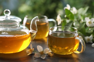 Photo of Tasty jasmine tea in cups, teapot and flowers on black table, closeup