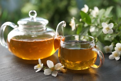 Photo of Tasty jasmine tea in cup, teapot and flowers on black table, closeup
