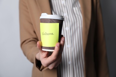 Photo of Welcome. Human resources manager holding takeaway cup with sticky note on grey background, closeup