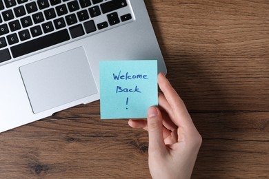 Photo of Woman holding paper note with words Welcome Back near laptop at wooden table, top view