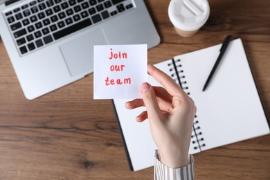 Photo of Woman holding paper note with words Join Our Team at wooden table, top view