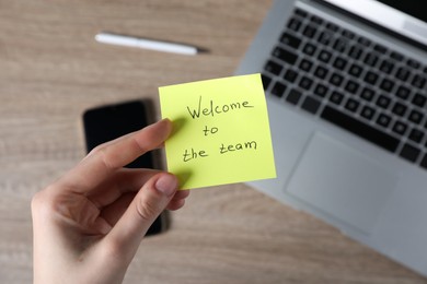 Photo of Woman holding paper note with words Welcome To The Team at wooden table, top view