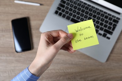 Photo of Woman holding paper note with words Welcome To The Team at wooden table, top view