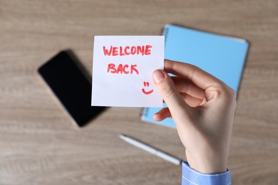 Photo of Woman holding paper note with words Welcome Back at wooden table, top view