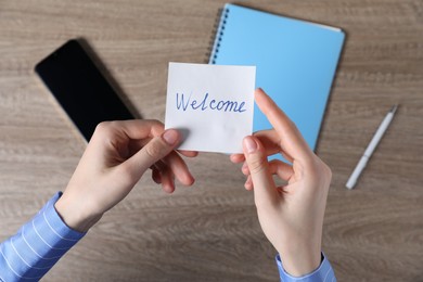 Photo of Woman holding paper note with word Welcome at wooden table, top view