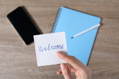 Photo of Woman holding paper note with word Welcome at wooden table, top view