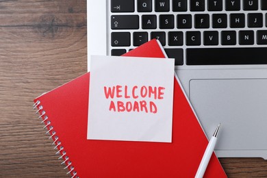 Photo of Paper note with words Welcome Aboard, notepad, laptop and pen on wooden table, flat lay