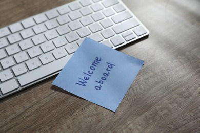 Photo of Paper note with words Welcome Aboard and computer keyboard on wooden table, closeup