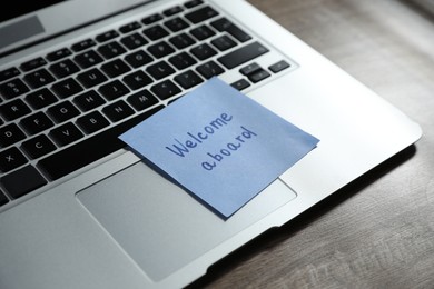 Photo of Paper note with words Welcome Aboard and laptop on wooden table, closeup