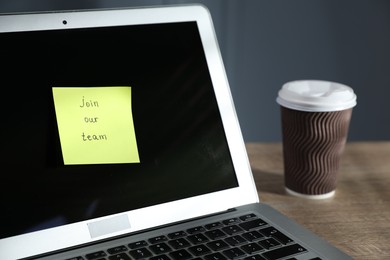 Photo of Sticky note with words Join Our Team, laptop and takeaway cup on wooden table against grey background, closeup