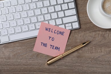 Photo of Paper note with words Welcome To The Team, pen and computer keyboard on wooden table, flat lay