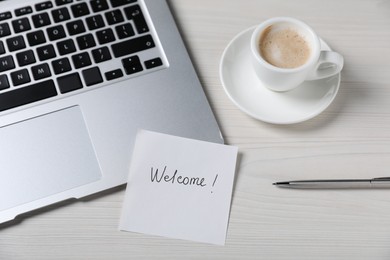 Photo of Paper note with word Welcome, laptop, cup of coffee and pen on light wooden table, above view