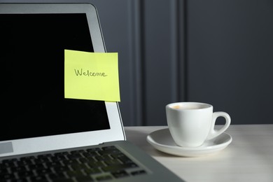 Photo of Sticky note with word Welcome, laptop and cup on white wooden table against grey background, closeup