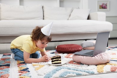 Photo of Birthday celebration. Little boy with piece of cake having video call via laptop at home