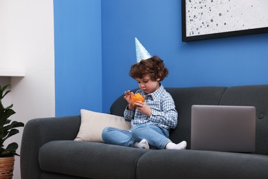 Photo of Birthday celebration. Little boy with cupcake having video call via laptop on sofa at home