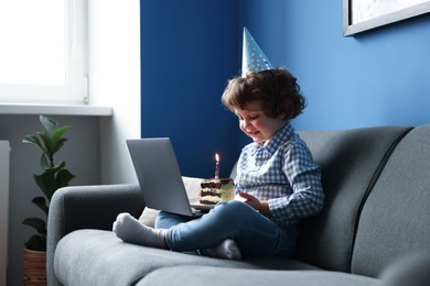 Photo of Birthday celebration. Little boy with piece of cake having video call via laptop on sofa at home