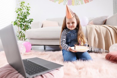 Photo of Birthday celebration. Little girl with piece of cake having video call via laptop at home