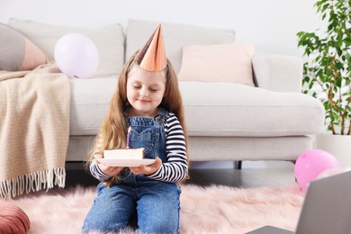 Photo of Birthday celebration. Little girl with piece of cake having video call via laptop at home