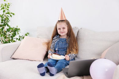 Photo of Birthday celebration. Little girl with piece of cake having video call via laptop on sofa at home