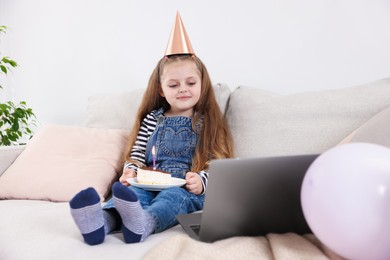 Photo of Birthday celebration. Little girl with piece of cake having video call via laptop on sofa at home