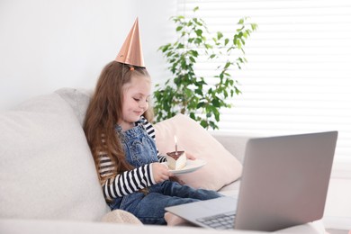 Photo of Birthday celebration. Little girl with piece of cake having video call via laptop on sofa at home