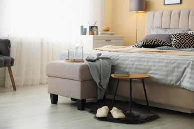 Photo of Comfortable bed with soft linens, slippers, pouf and side table in room. Home textile