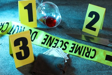 Photo of Forensic expert. Yellow crime scene tape, markers, wallet with money, knife and glass of wine on grey table, closeup