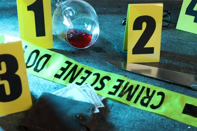 Photo of Forensic expert. Yellow crime scene tape, markers, wallet with money, knife and glass of wine on grey table, closeup