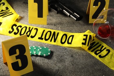 Photo of Forensic expert. Yellow crime scene tape, markers, poker chips, gun and glass of wine on grey table, closeup