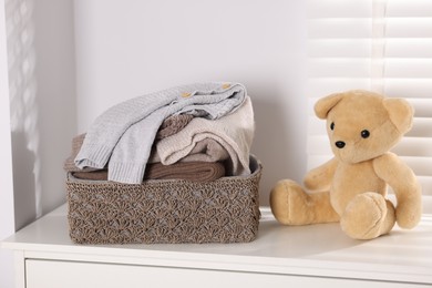 Photo of Wool sweaters in basket and teddy bear on white nightstand indoors