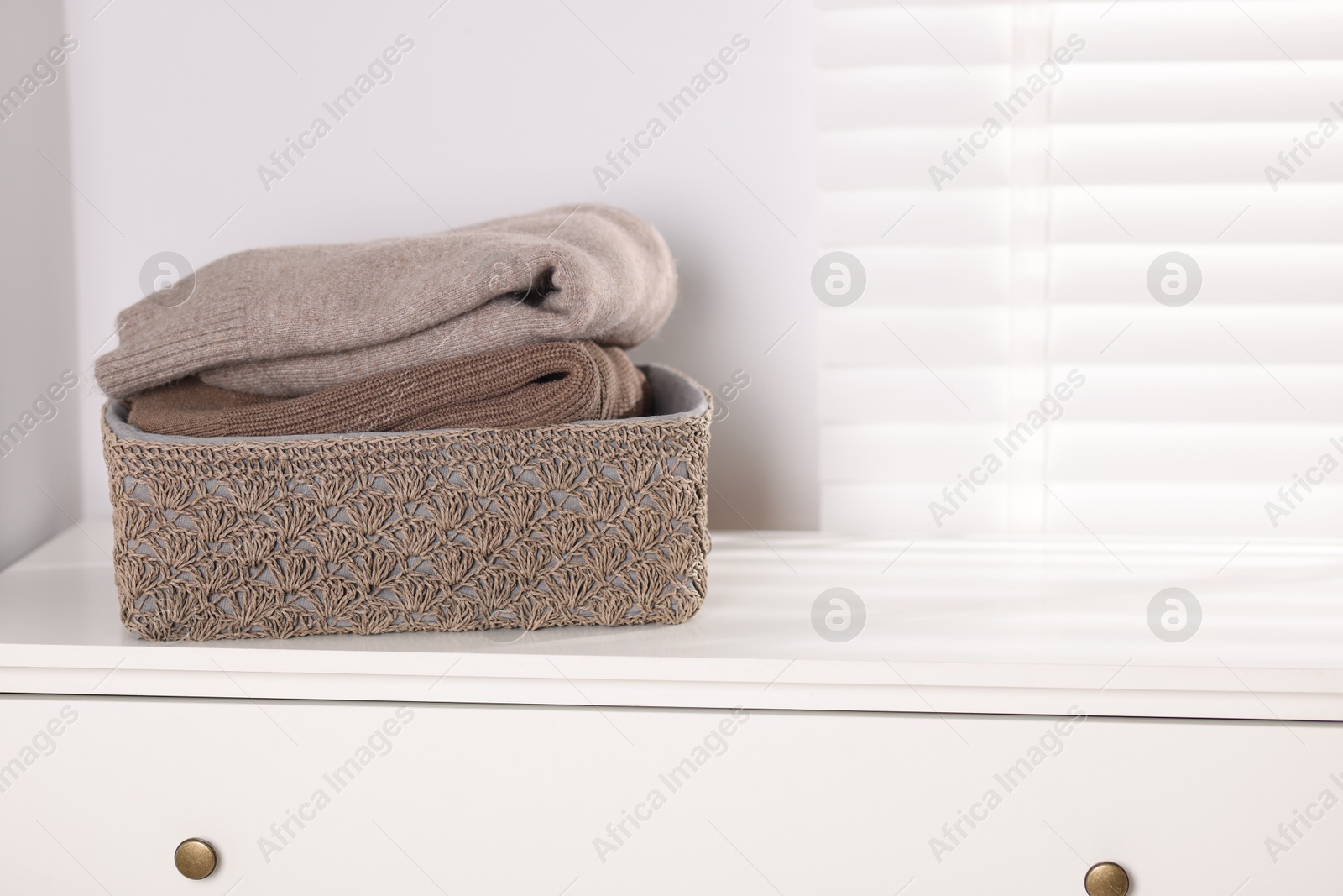 Wool sweaters in basket on white nightstand indoors, space for text Photo of Wool sweaters in basket on white nightstand indoors, space for text