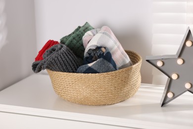 Photo of Wool scarves in wicker basket and star shaped lamp on white nightstand indoors, closeup