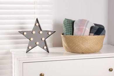 Photo of Wool scarves in wicker basket and star shaped lamp on white nightstand indoors