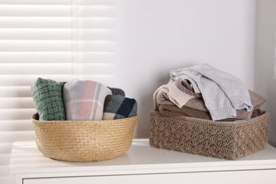 Photo of Wool sweaters and scarves in baskets on white nightstand indoors