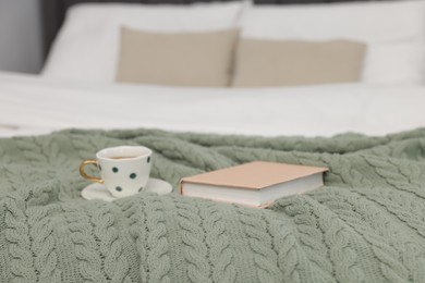 Photo of Woolen blanket, cup of drink and book on bed indoors, closeup