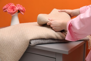 Photo of Woman folding woolen blanket near orange wall indoors, closeup