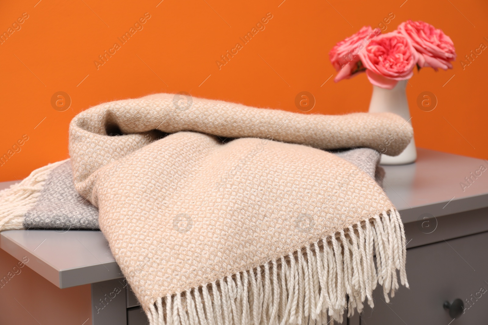 Woolen blankets and flowers on cabinet near orange wall indoors, closeup Photo of Woolen blankets and flowers on cabinet near orange wall indoors, closeup
