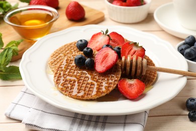 Photo of Tasty Dutch waffles (stroopwafels) with honey, strawberries and blueberries on light wooden table, closeup