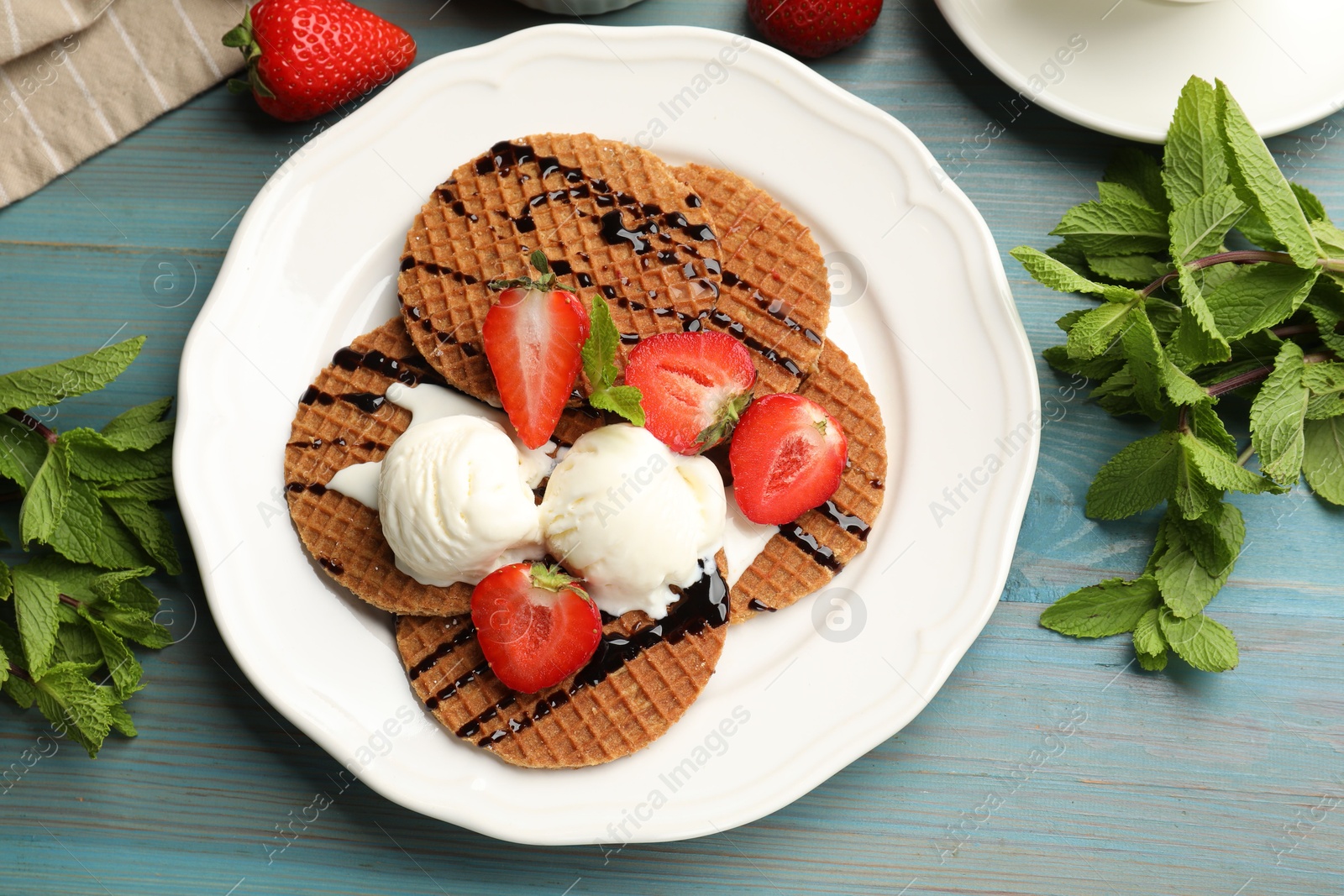 Tasty Dutch waffles (stroopwafels) with ice cream, chocolate sauce, mint and strawberries on light blue wooden table, flat lay Photo of Tasty Dutch waffles (stroopwafels) with ice cream, chocolate sauce, mint and strawberries on light blue wooden table, flat lay