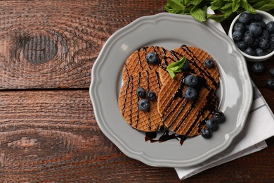 Photo of Tasty Dutch waffles (stroopwafels) with blueberries, mint and chocolate sauce on wooden table, flat lay. Space for text