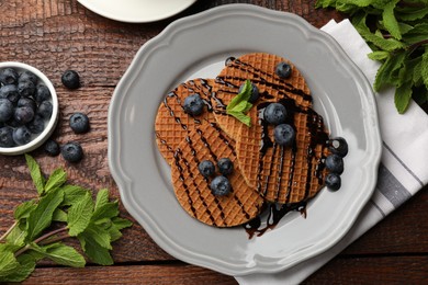 Photo of Tasty Dutch waffles (stroopwafels) with blueberries, mint and chocolate sauce on wooden table, flat lay
