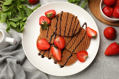 Photo of Tasty Dutch waffles (stroopwafels) with strawberries, mint and chocolate sauce on grey textured table, flat lay